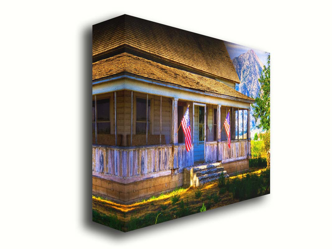 A photo of a weatherd home, with two American flags hanging from the porch. Green plants are growing in the yard, and a mountain can be seen in the background. Printed on canvas.