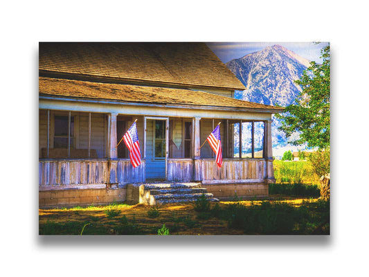 A photo of a weatherd home, with two American flags hanging from the porch. Green plants are growing in the yard, and a mountain can be seen in the background. Printed on canvas.