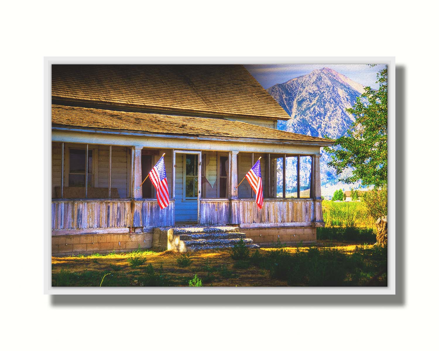 A photo of a weatherd home, with two American flags hanging from the porch. Green plants are growing in the yard, and a mountain can be seen in the background. Printed on canvas in a float frame.