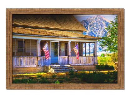 A photo of a weatherd home, with two American flags hanging from the porch. Green plants are growing in the yard, and a mountain can be seen in the background. Printed on canvas and framed.