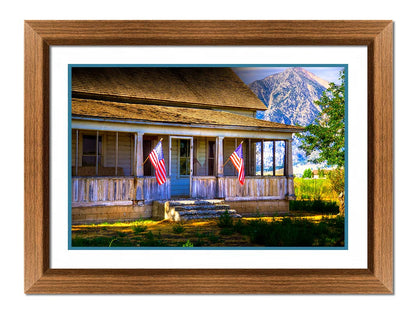 A photo of a weatherd home, with two American flags hanging from the porch. Green plants are growing in the yard, and a mountain can be seen in the background. Printed on paper, matted, and framed.