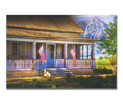 A photo of a weatherd home, with two American flags hanging from the porch. Green plants are growing in the yard, and a mountain can be seen in the background. Printed on a box board.