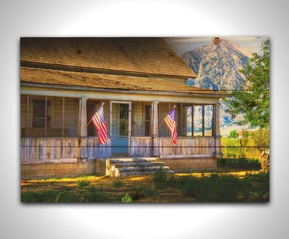 A photo of a weatherd home, with two American flags hanging from the porch. Green plants are growing in the yard, and a mountain can be seen in the background. Printed on a wood pallet.