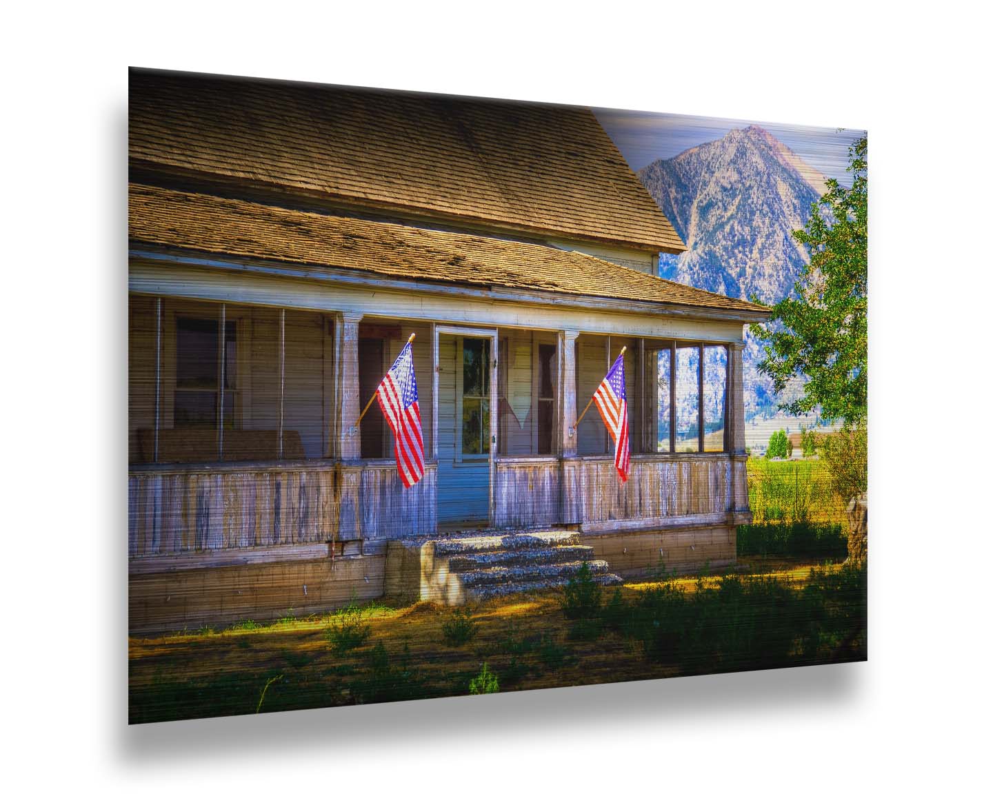 A photo of a weatherd home, with two American flags hanging from the porch. Green plants are growing in the yard, and a mountain can be seen in the background. Printed on metal.