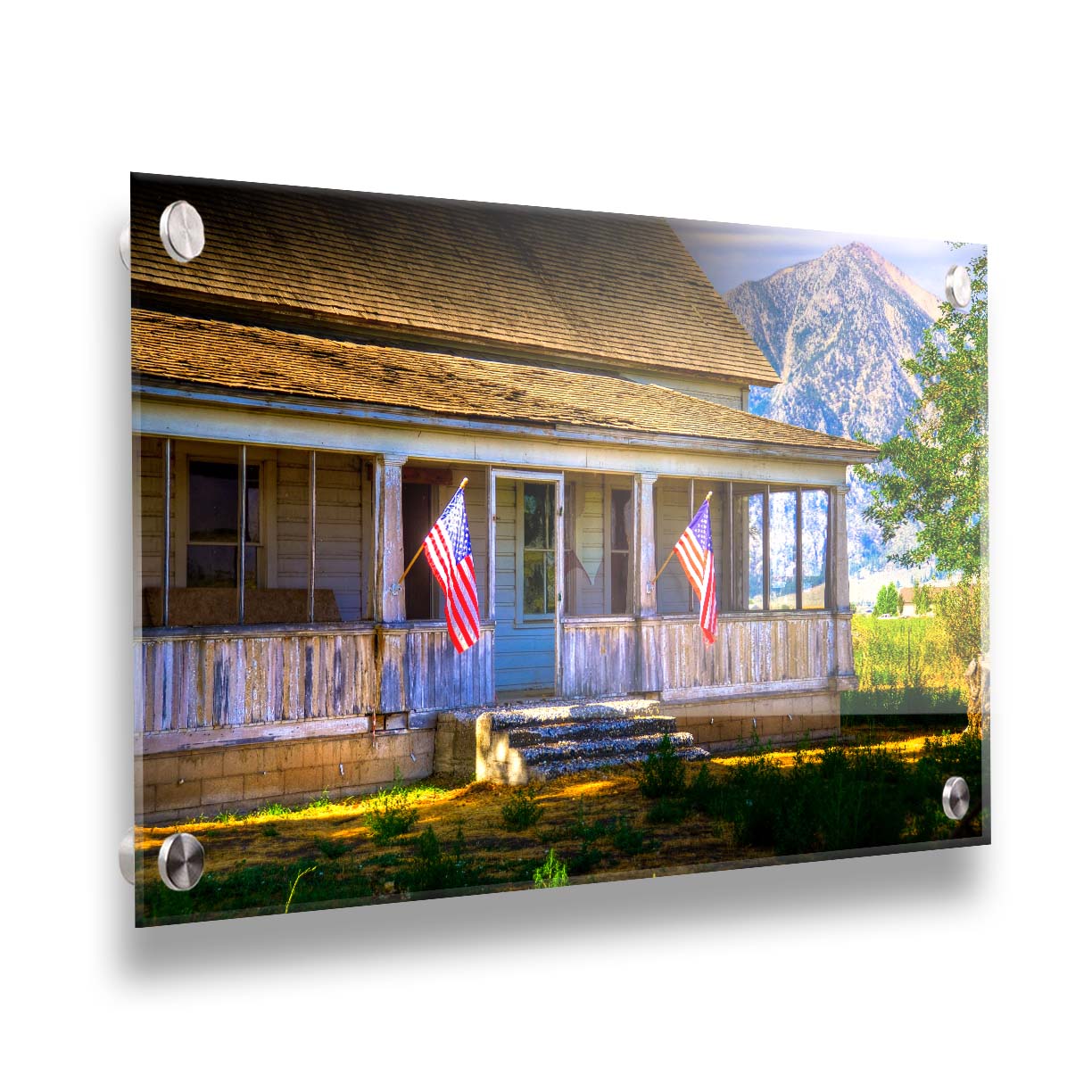 A photo of a weatherd home, with two American flags hanging from the porch. Green plants are growing in the yard, and a mountain can be seen in the background. Printed on acrylic.