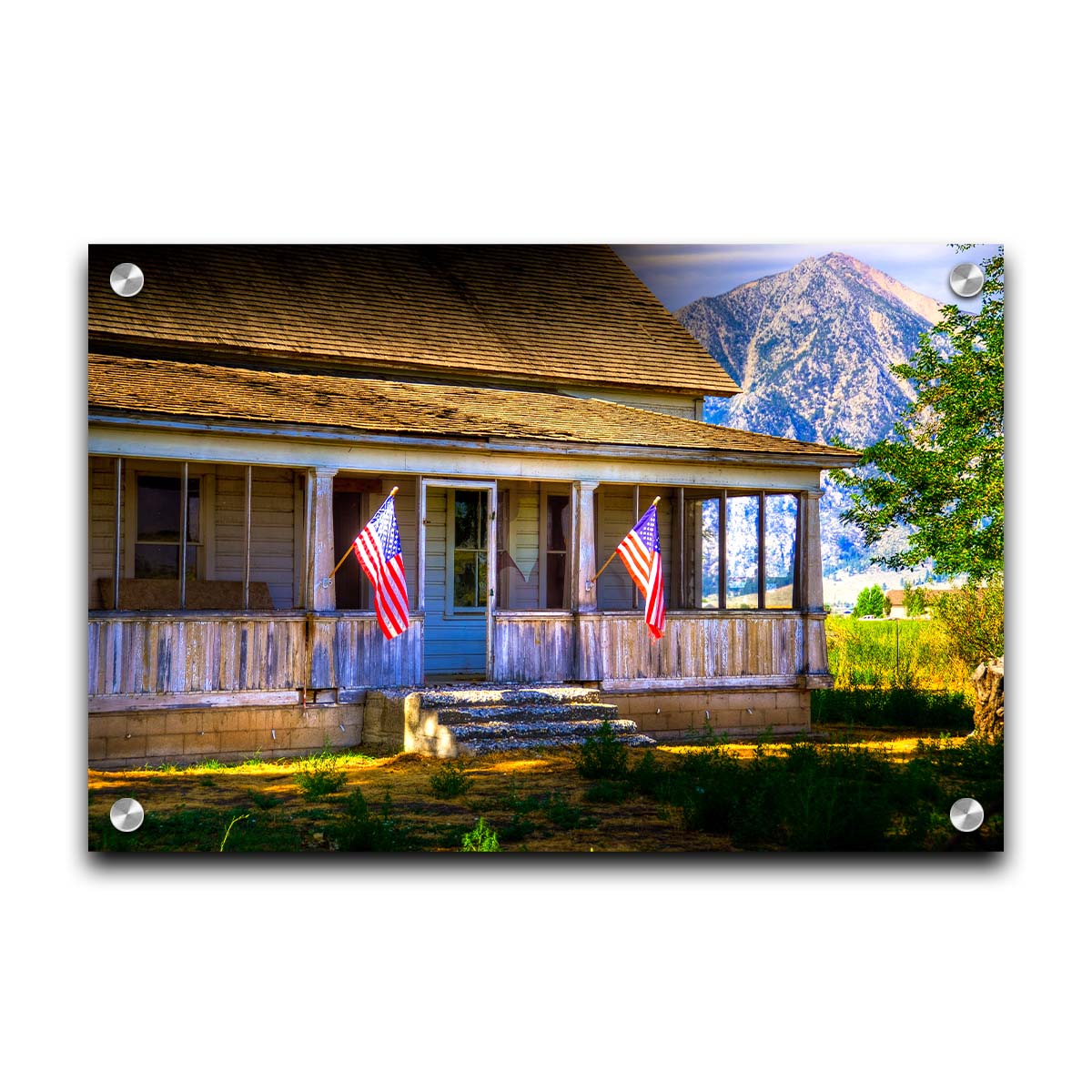 A photo of a weatherd home, with two American flags hanging from the porch. Green plants are growing in the yard, and a mountain can be seen in the background. Printed on acrylic.