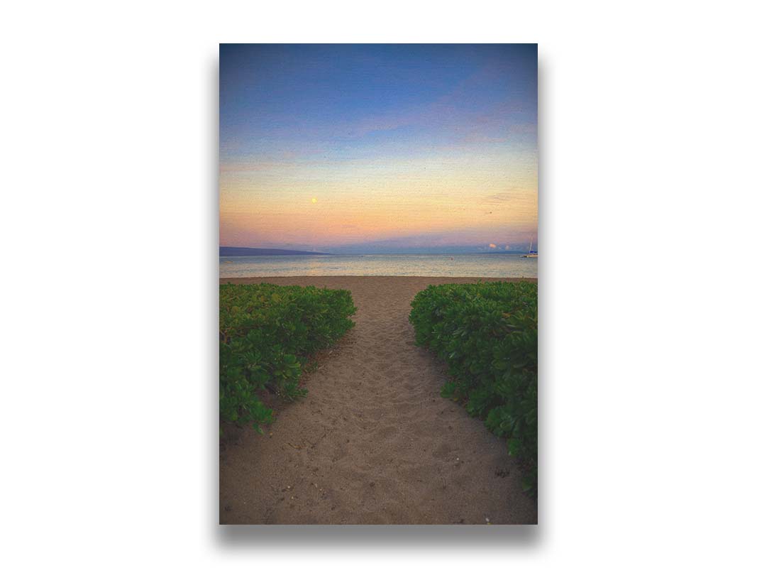 A photo looking out to sea at Kaʻanapali beach at sunset. Lush foliage lines the path down the beach, and sail boats can be see on the water. Printed on canvas.