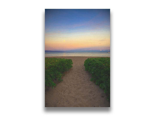 A photo looking out to sea at Kaʻanapali beach at sunset. Lush foliage lines the path down the beach, and sail boats can be see on the water. Printed on canvas.