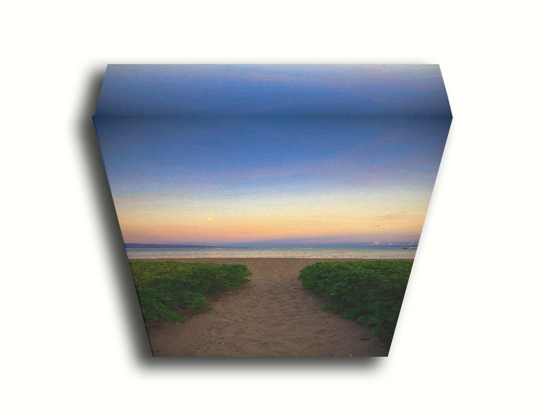 A photo looking out to sea at Kaʻanapali beach at sunset. Lush foliage lines the path down the beach, and sail boats can be see on the water. Printed on canvas.