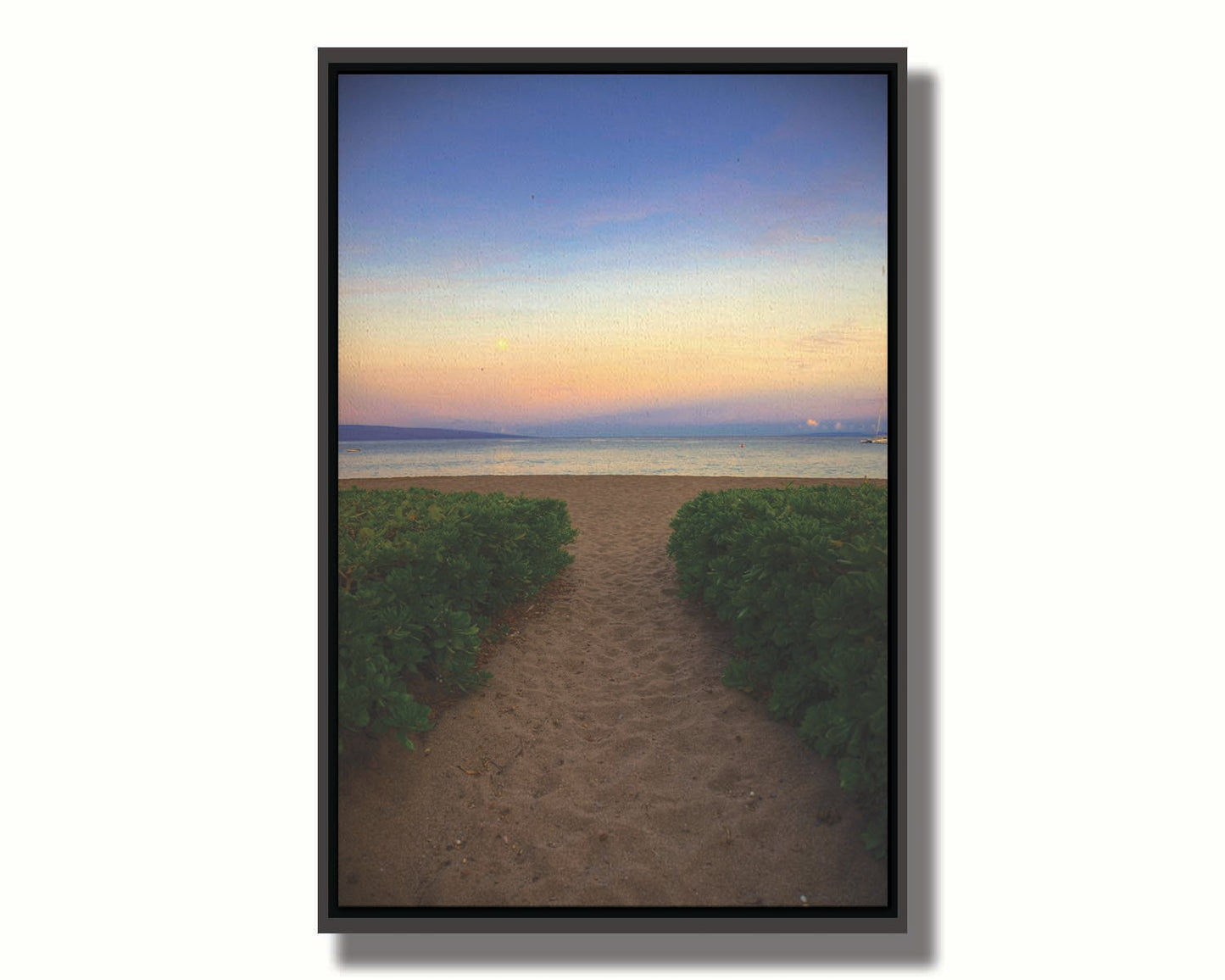 A photo looking out to sea at Kaʻanapali beach at sunset. Lush foliage lines the path down the beach, and sail boats can be see on the water. Printed on canvas in a float frame.