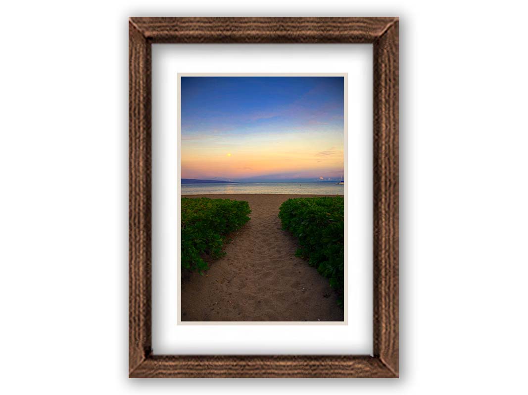 A photo looking out to sea at Kaʻanapali beach at sunset. Lush foliage lines the path down the beach, and sail boats can be see on the water. Printed on paper, matted, and framed.