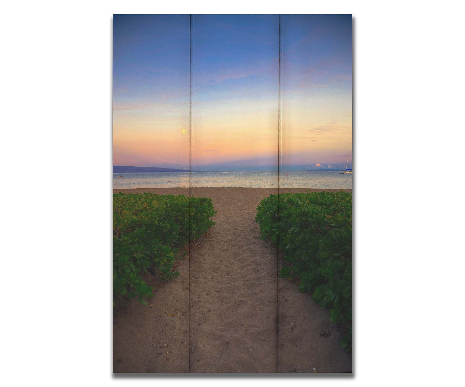 A photo looking out to sea at Kaʻanapali beach at sunset. Lush foliage lines the path down the beach, and sail boats can be see on the water. Printed on a box board.