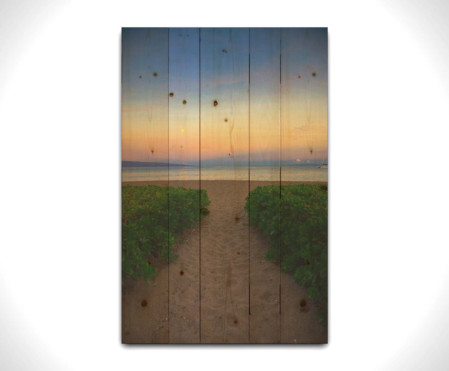 A photo looking out to sea at Kaʻanapali beach at sunset. Lush foliage lines the path down the beach, and sail boats can be see on the water. Printed on a wood pallet.