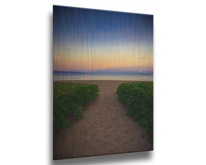 A photo looking out to sea at Kaʻanapali beach at sunset. Lush foliage lines the path down the beach, and sail boats can be see on the water. Printed on metal.