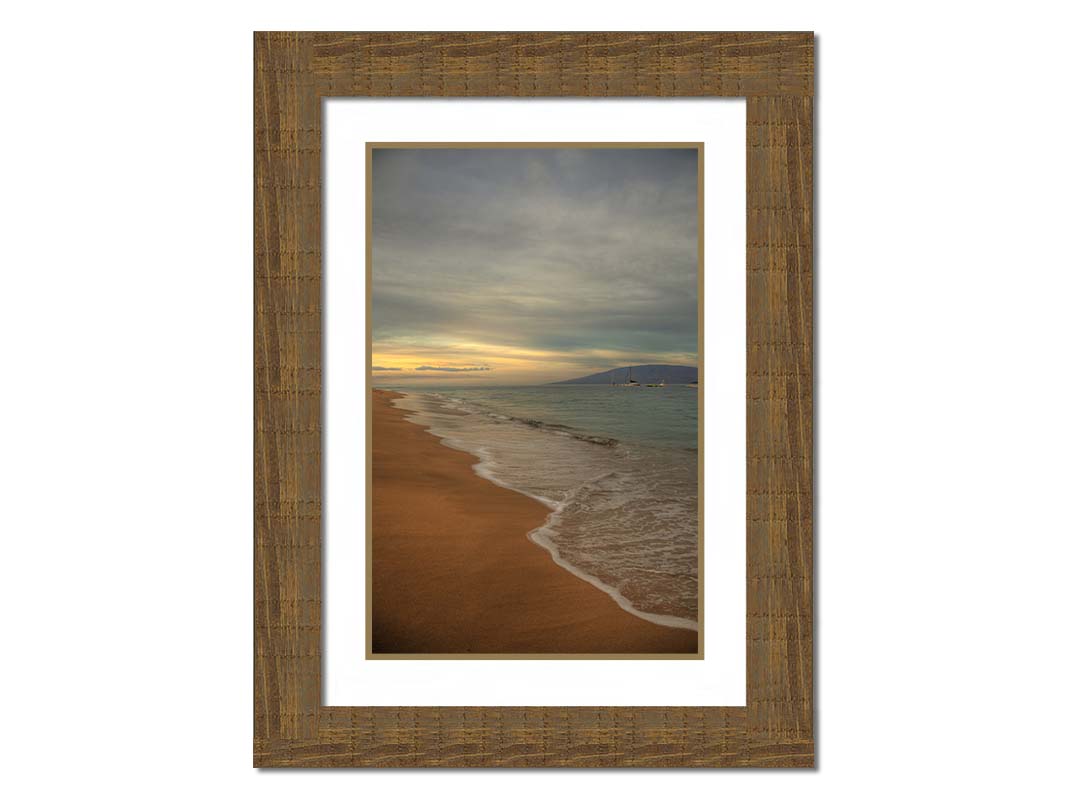 A photo of a cloudy sunrise on the Kaʻanapali shore, looking out to sea. Sailboats can be seen out on the water. Printed on paper, matted, and framed.