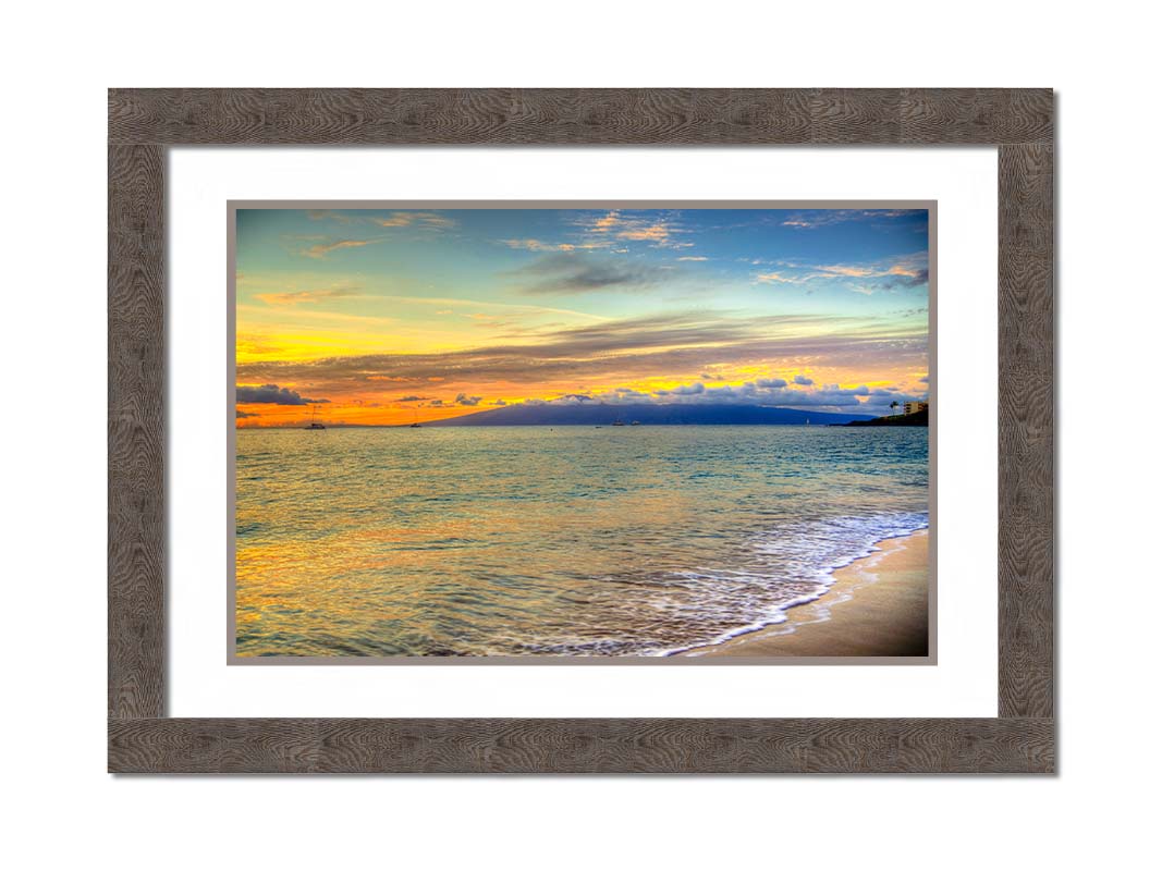 A photo of the sunset on the beach at Kaʻanapali, looking out to sea. Sailboats can be seen on the water, and mountains stand tall among clouds in the background. Printed on paper, matted, and framed.