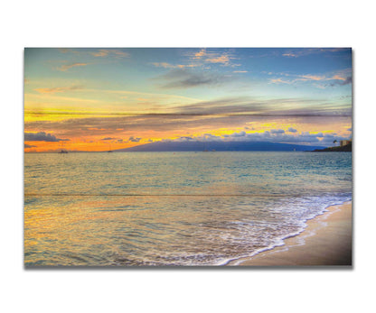 A photo of the sunset on the beach at Kaʻanapali, looking out to sea. Sailboats can be seen on the water, and mountains stand tall among clouds in the background. Printed on a box board.