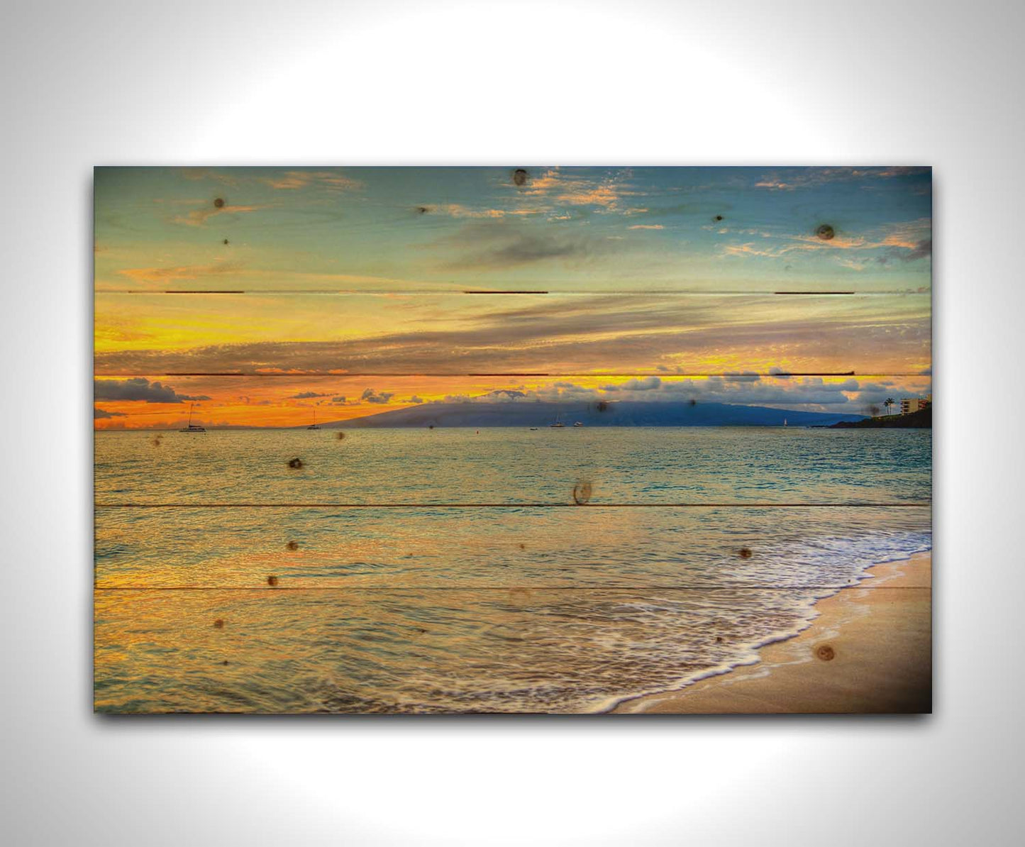 A photo of the sunset on the beach at Kaʻanapali, looking out to sea. Sailboats can be seen on the water, and mountains stand tall among clouds in the background. Printed on a wood pallet.