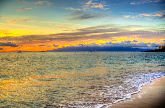 A photo of the sunset on the beach at Kaʻanapali, looking out to sea. Sailboats can be seen on the water, and mountains stand tall among clouds in the background.