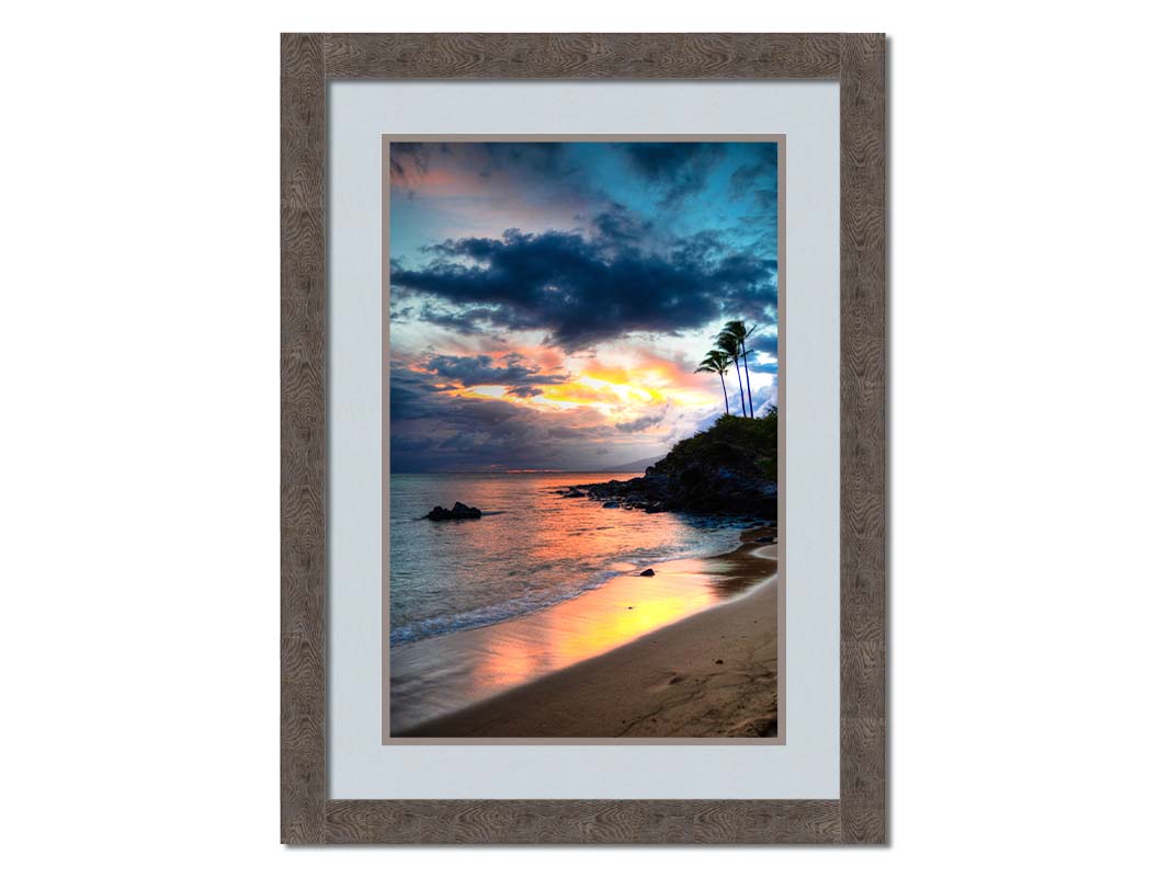 A photo of the sunset at Honokahua, Maui peeking through the clouds and reflecting pink and orange on the water. Palms are silhouetted against the sky. Printed on paper, matted, and framed.