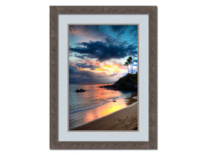 A photo of the sunset at Honokahua, Maui peeking through the clouds and reflecting pink and orange on the water. Palms are silhouetted against the sky. Printed on paper, matted, and framed.