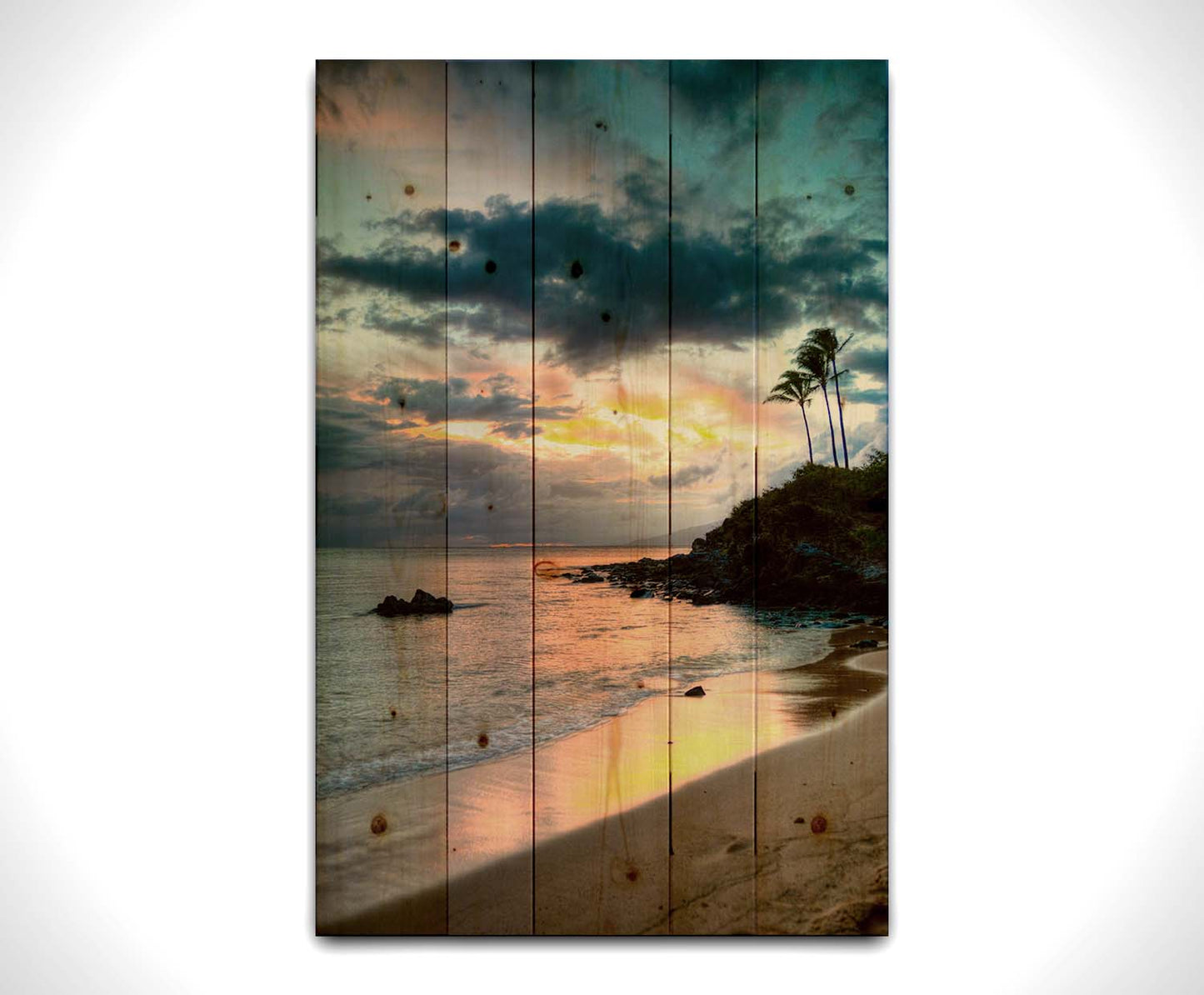 A photo of the sunset at Honokahua, Maui peeking through the clouds and reflecting pink and orange on the water. Palms are silhouetted against the sky. Printed on a wood pallet.