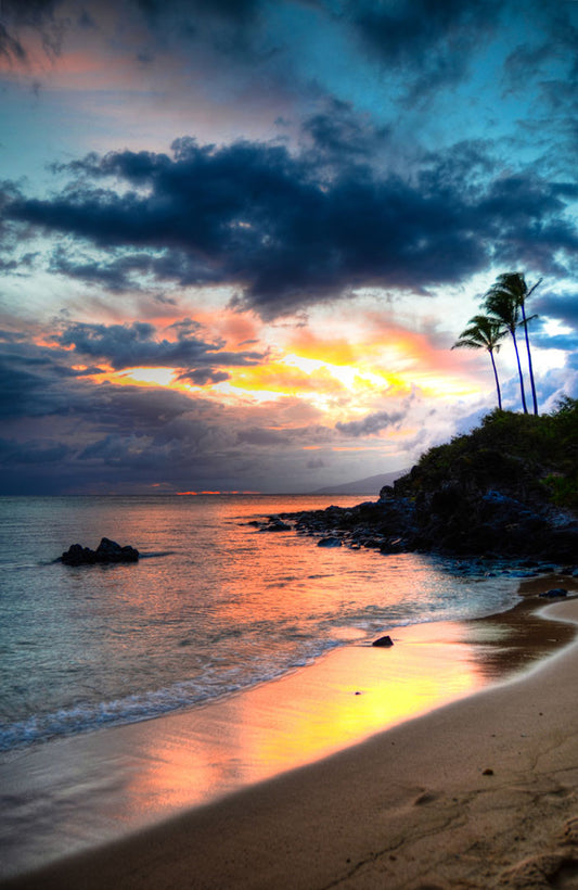 A photo of the sunset at Honokahua, Maui peeking through the clouds and reflecting pink and orange on the water. Palms are silhouetted against the sky. 