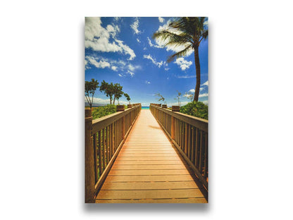 A photo of a boardwalk in Maui, Hawaiʻi, looking down the wood path to the sea. It is flanked by shrubbery, palms, and other tropical trees. Printed on canvas.