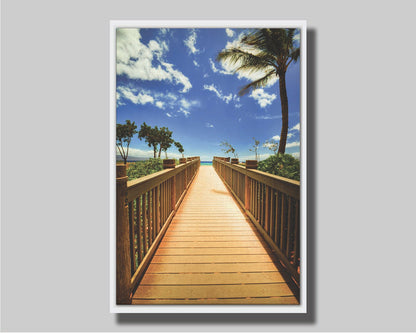 A photo of a boardwalk in Maui, Hawaiʻi, looking down the wood path to the sea. It is flanked by shrubbery, palms, and other tropical trees. Printed on canvas in a float frame.