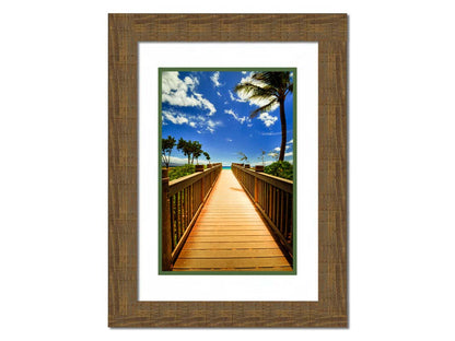 A photo of a boardwalk in Maui, Hawaiʻi, looking down the wood path to the sea. It is flanked by shrubbery, palms, and other tropical trees. Printed on paper, matted, and framed.