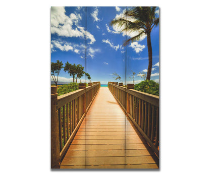 A photo of a boardwalk in Maui, Hawaiʻi, looking down the wood path to the sea. It is flanked by shrubbery, palms, and other tropical trees. Printed on a box board.