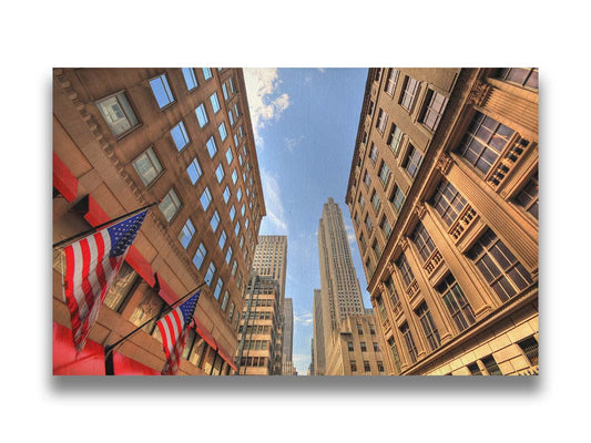 A photo looking up along a New York street lined with buildings. American flags hang on poles off the side of one of the buildings. Printed on canvas.