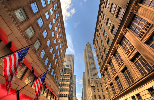 A photo looking up along a New York street lined with buildings. American flags hang on poles off the side of one of the buildings.