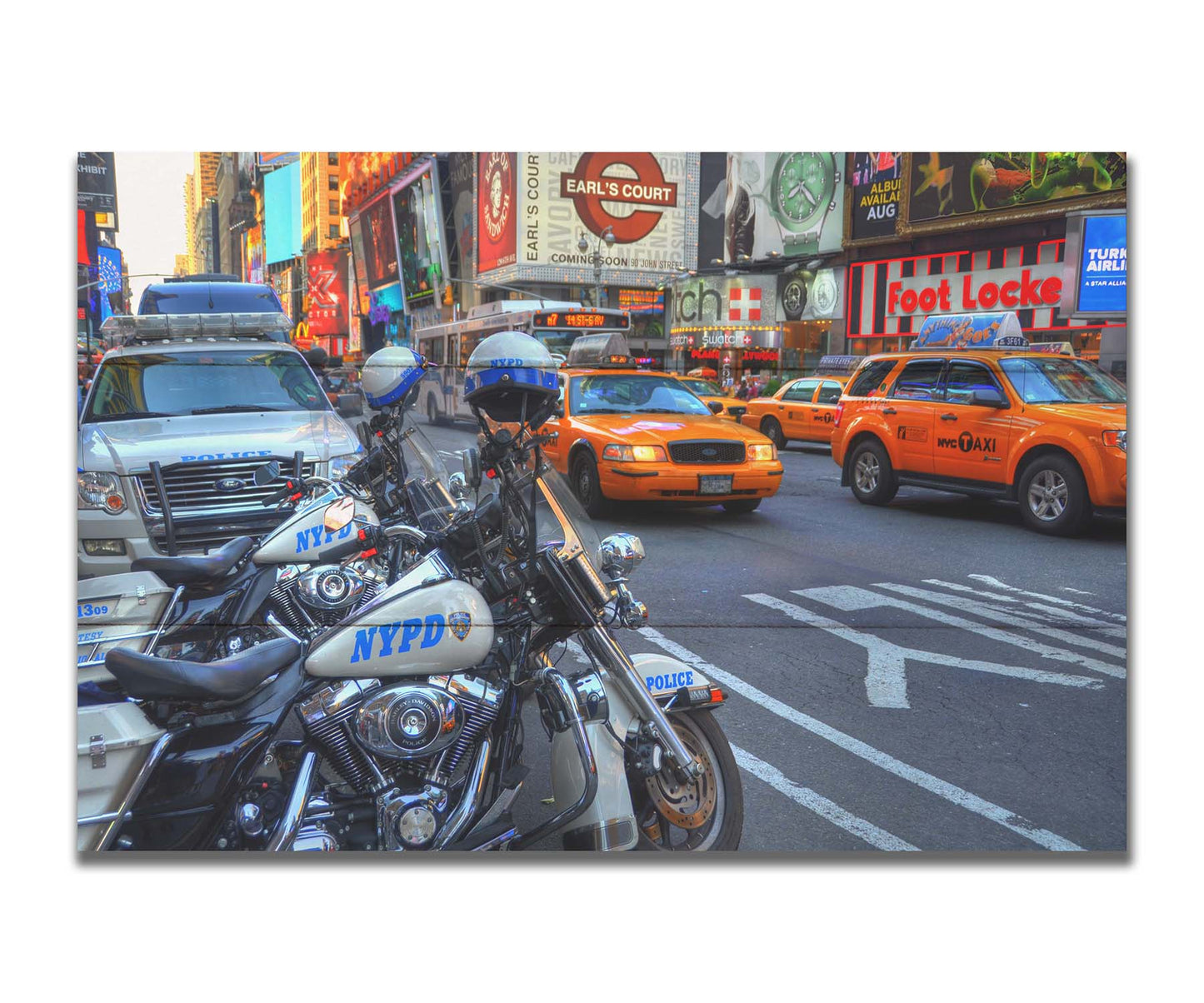 A photo of a street in New York City, featuring several bright orange taxis and New York Police Department vehicles. Printed on a box board.