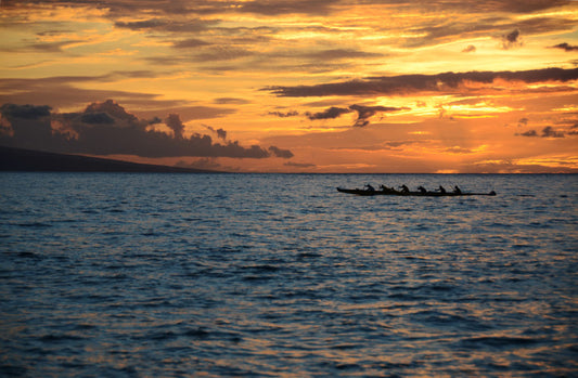 A photo of an outrigger canoe silhouetted on the sea by an orange sunset off the coat of Hawaiʻi.