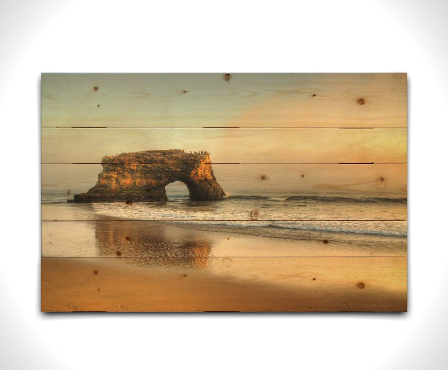 A photo of a "natural bridge" rock formation in Santa Cruz, California. A group of pelicans sit on top of the structure above the sea. Printed on a wood pallet.