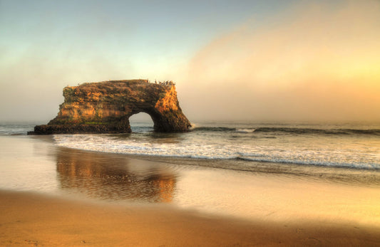 A photo of a "natural bridge" rock formation in Santa Cruz, California. A group of pelicans sit on top of the structure above the sea.