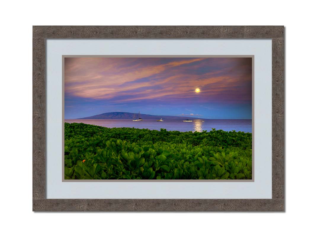 A photo overlooking the sea off the coast of Hawaiʻi as the moon sets. The foreground is filled with greenery, and ships can be seen sailing on the waters with mountains in the distance. Printed on paper, matted, and framed.