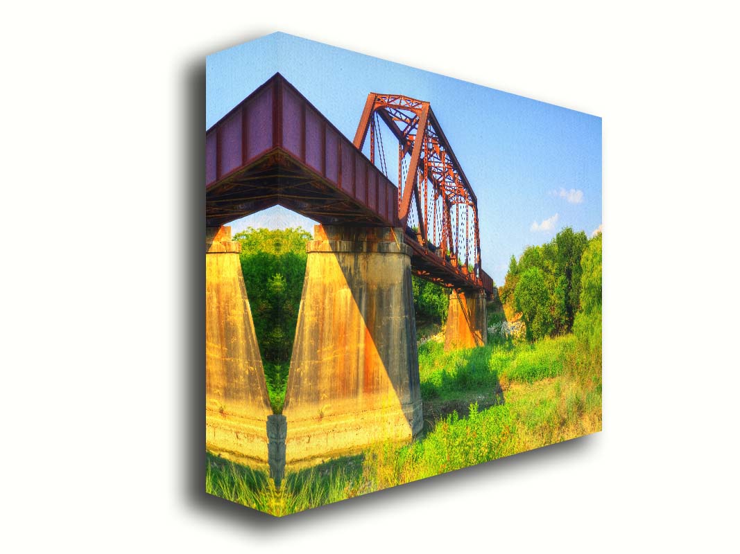 A photograph of a red metal bridge supported by concrete pillars, passing ove a grassy landscape on a clear day. Printed on canvas.