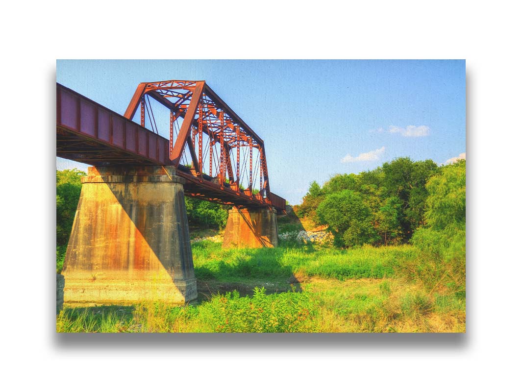 A photograph of a red metal bridge supported by concrete pillars, passing ove a grassy landscape on a clear day. Printed on canvas.