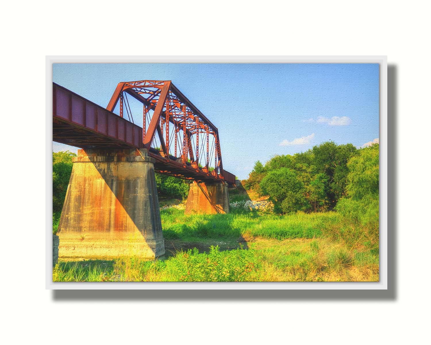 A photograph of a red metal bridge supported by concrete pillars, passing ove a grassy landscape on a clear day. Printed on canvas in a float frame.