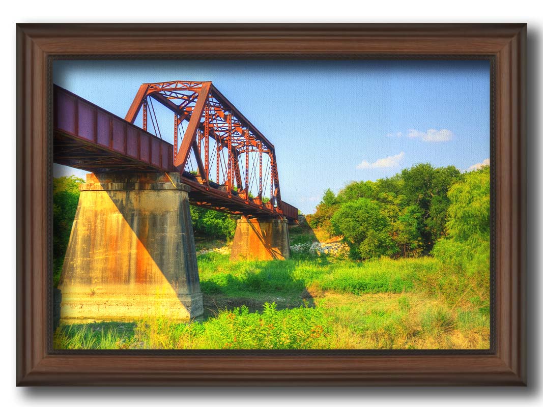 A photograph of a red metal bridge supported by concrete pillars, passing ove a grassy landscape on a clear day. Printed on canvas and framed.