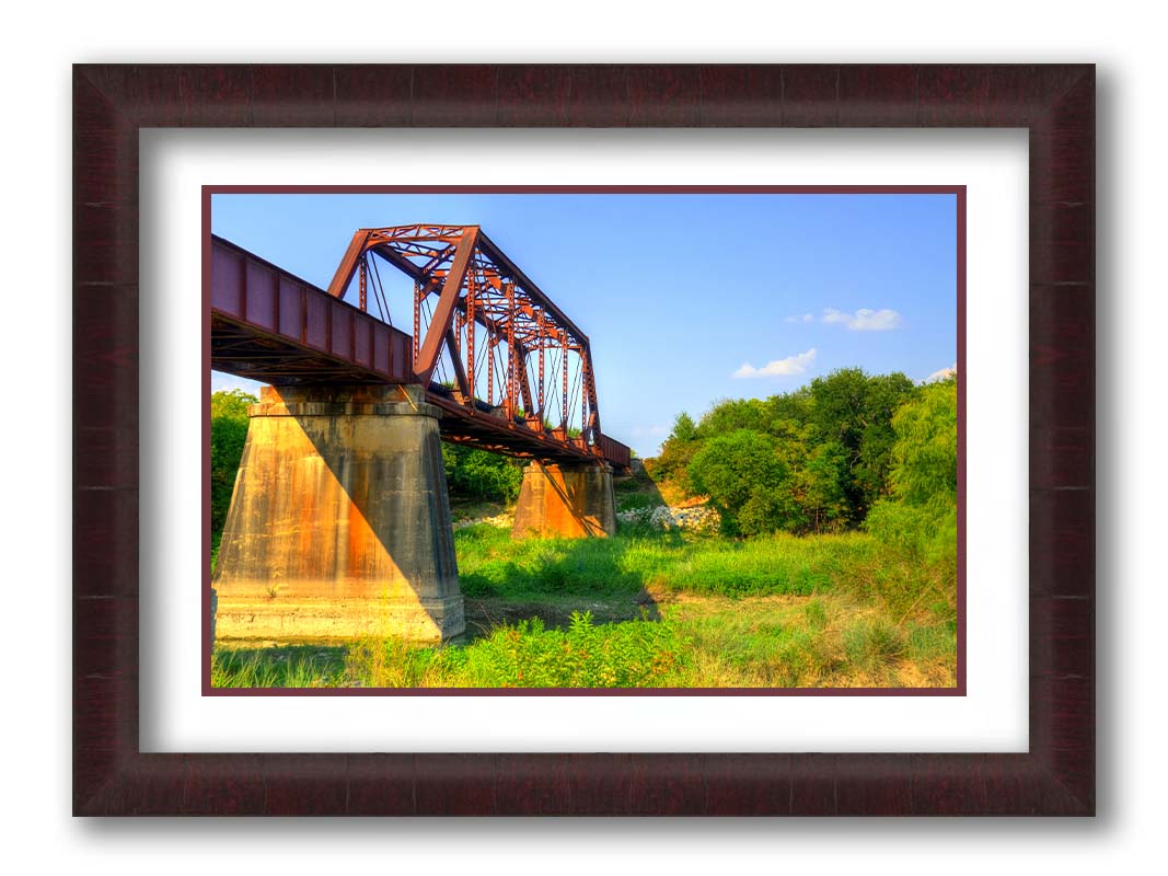 A photograph of a red metal bridge supported by concrete pillars, passing ove a grassy landscape on a clear day. Printed on paper, matted, and framed.