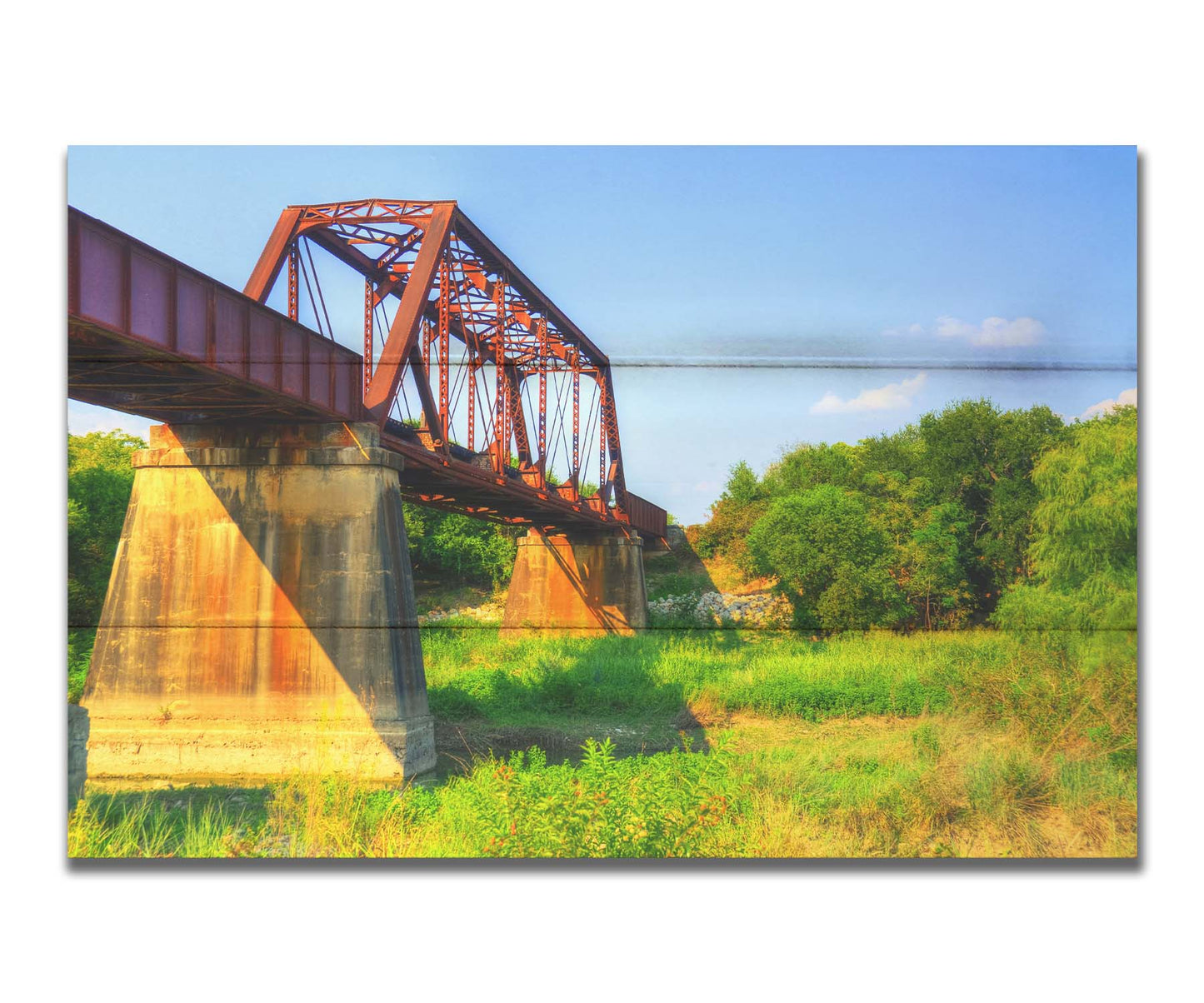 A photograph of a red metal bridge supported by concrete pillars, passing ove a grassy landscape on a clear day. Printed on a box board.