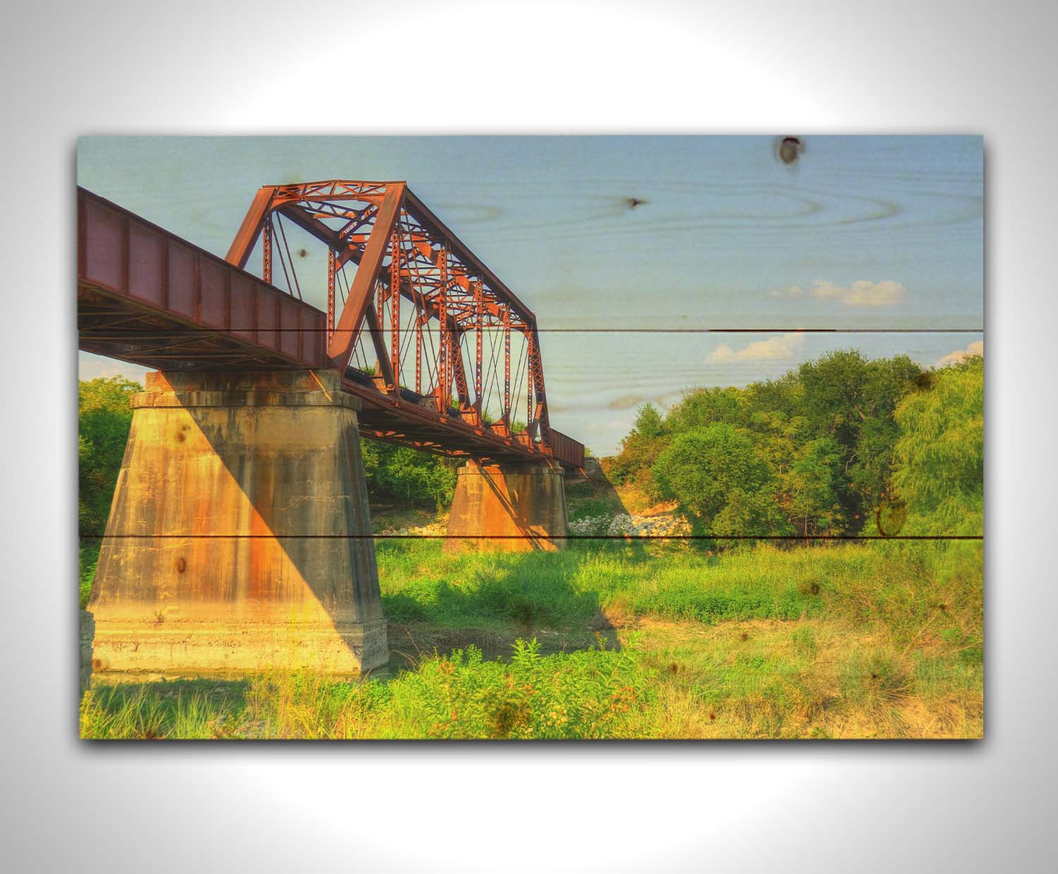 A photograph of a red metal bridge supported by concrete pillars, passing ove a grassy landscape on a clear day. Printed on a wood pallet.