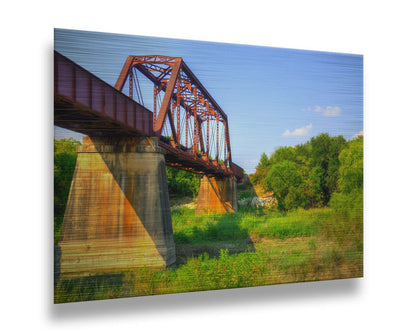 A photograph of a red metal bridge supported by concrete pillars, passing ove a grassy landscape on a clear day. Printed on metal.