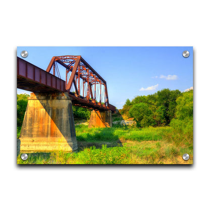 A photograph of a red metal bridge supported by concrete pillars, passing ove a grassy landscape on a clear day. Printed on acrylic.