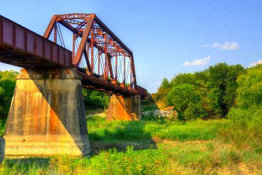 A photograph of a red metal bridge supported by concrete pillars, passing over a grassy landscape on a clear day.