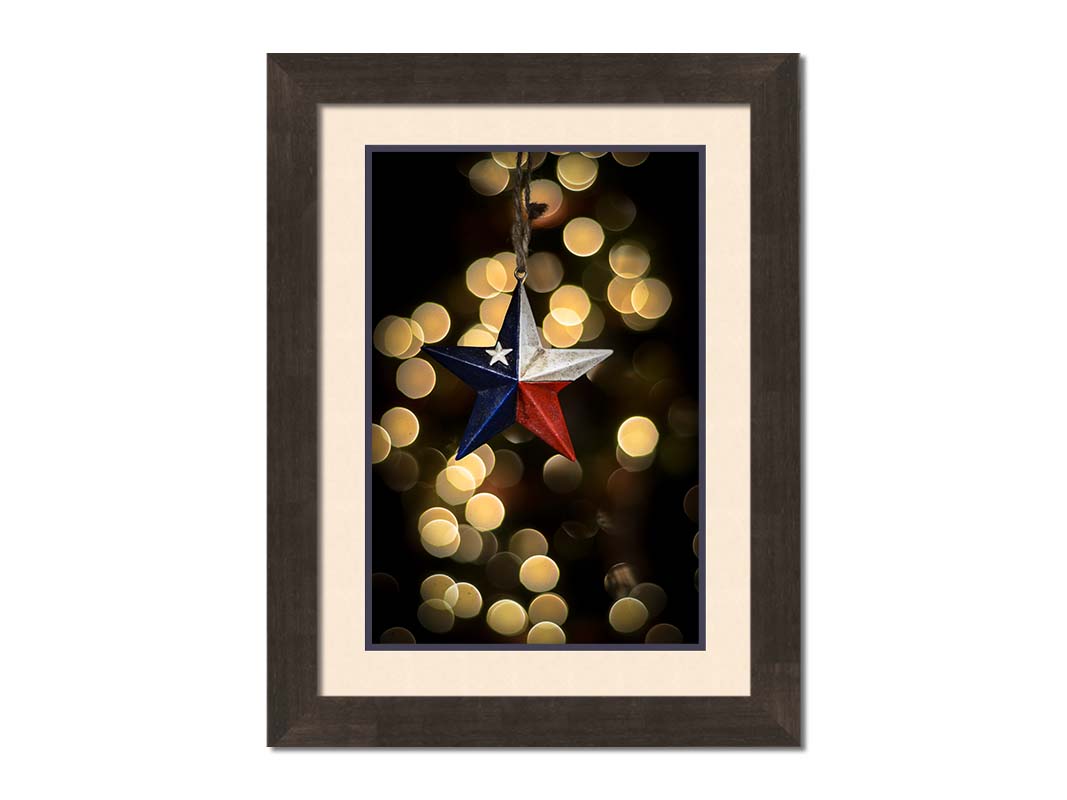 A photo of a christmas ornament of a star painted like the Texas state flag, with yellow bokeh in the background. Printed on paper, matted, and framed.