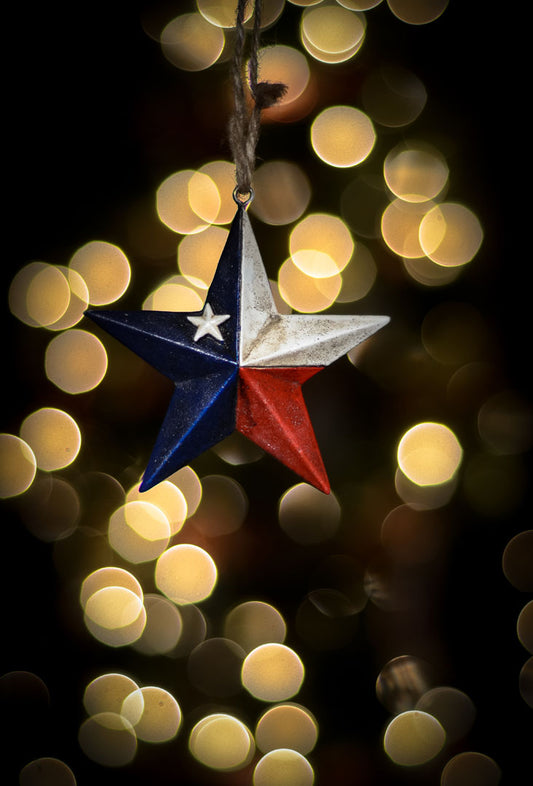 A photo of a christmas ornament of a star painted like the Texas state flag, with yellow bokeh in the background. 